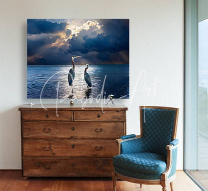 A framed canvas print titled Blue Herons At Dawn showing two herons in water under a dramatic cloudy sky with sunlight
