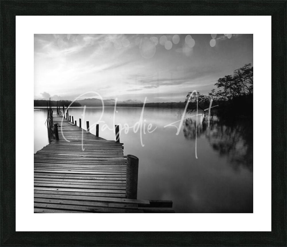 Black-framed black and white photographic print of a wooden pier over calm water titled dawn breaks over still waters
