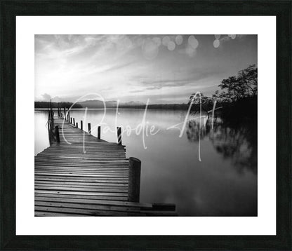 Black-framed black and white photographic print of a wooden pier over calm water titled dawn breaks over still waters