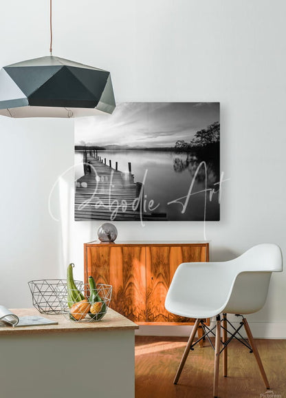 Black-and-white canvas print of wooden pier over calm water framed by trees titled dawn breaks over still waters