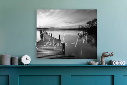 Black-and-white canvas print of wooden pier over calm water with trees and mountains titled dawn breaks over still waters