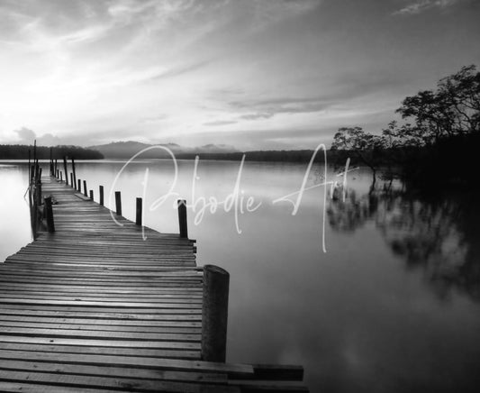 A weathered wooden dock extends into calm water in a monochrome scene titled dawn breaks over still waters