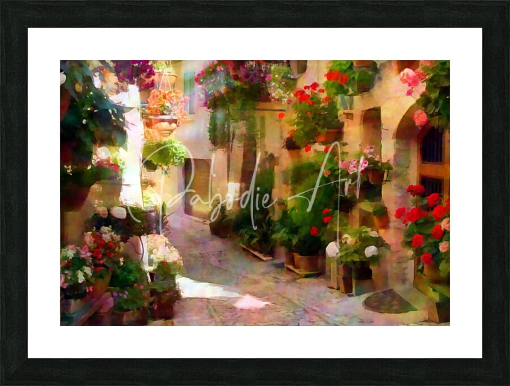 Black-framed art print of a sunlit cobblestone alley with vibrant red pink and white flowers titled flower filled cobblestone alley