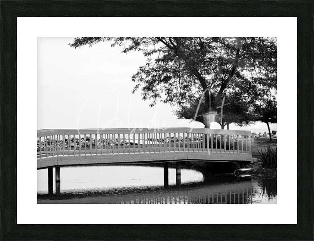 Black-framed black and white photo of sawmill creek wooden bridge with ornate railings over calm water and trees