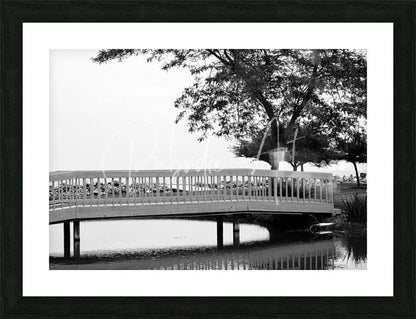 Black-framed black and white photo of sawmill creek wooden bridge with ornate railings over calm water and trees