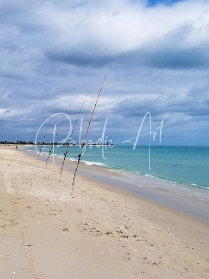 Three slender dark-colored fishing rods standing upright in sand near shoreline for shorefishing use